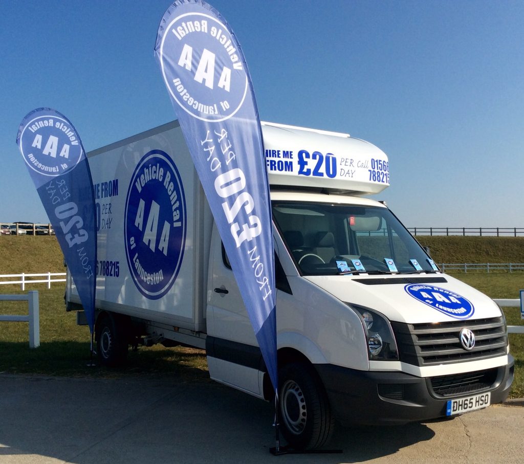 A white van from "AA Vehicle Rental" is parked outdoors, adorned with promotional flags and a handy price list on display. The signs proudly advertise rentals starting at just £20 per day. Branded with company logos, the scene unfolds under a clear blue sky.